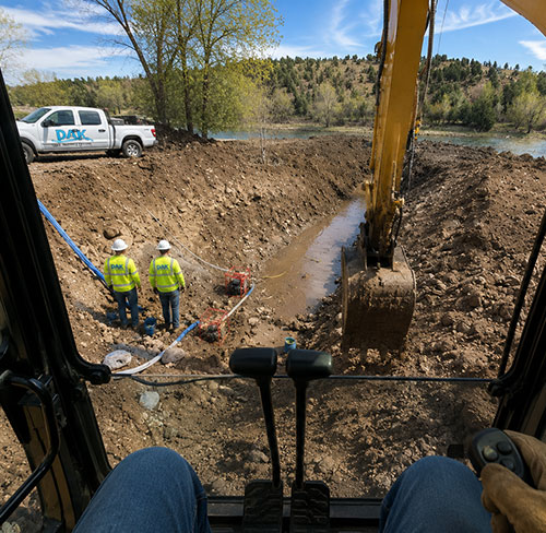 Excavator operator's view during gallery well construction near a river in Colorado, with DAK Drilling crew members installing groundwater collection infrastructure and excavation equipment on site.